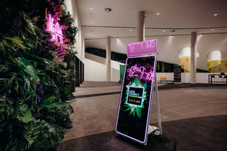 mirror photo booth in front of a tropical greenery wall with a pink let's party neon sign on it