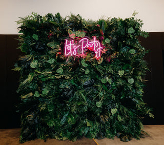 tropical greenery flower wall with pink let's party neon sign on it